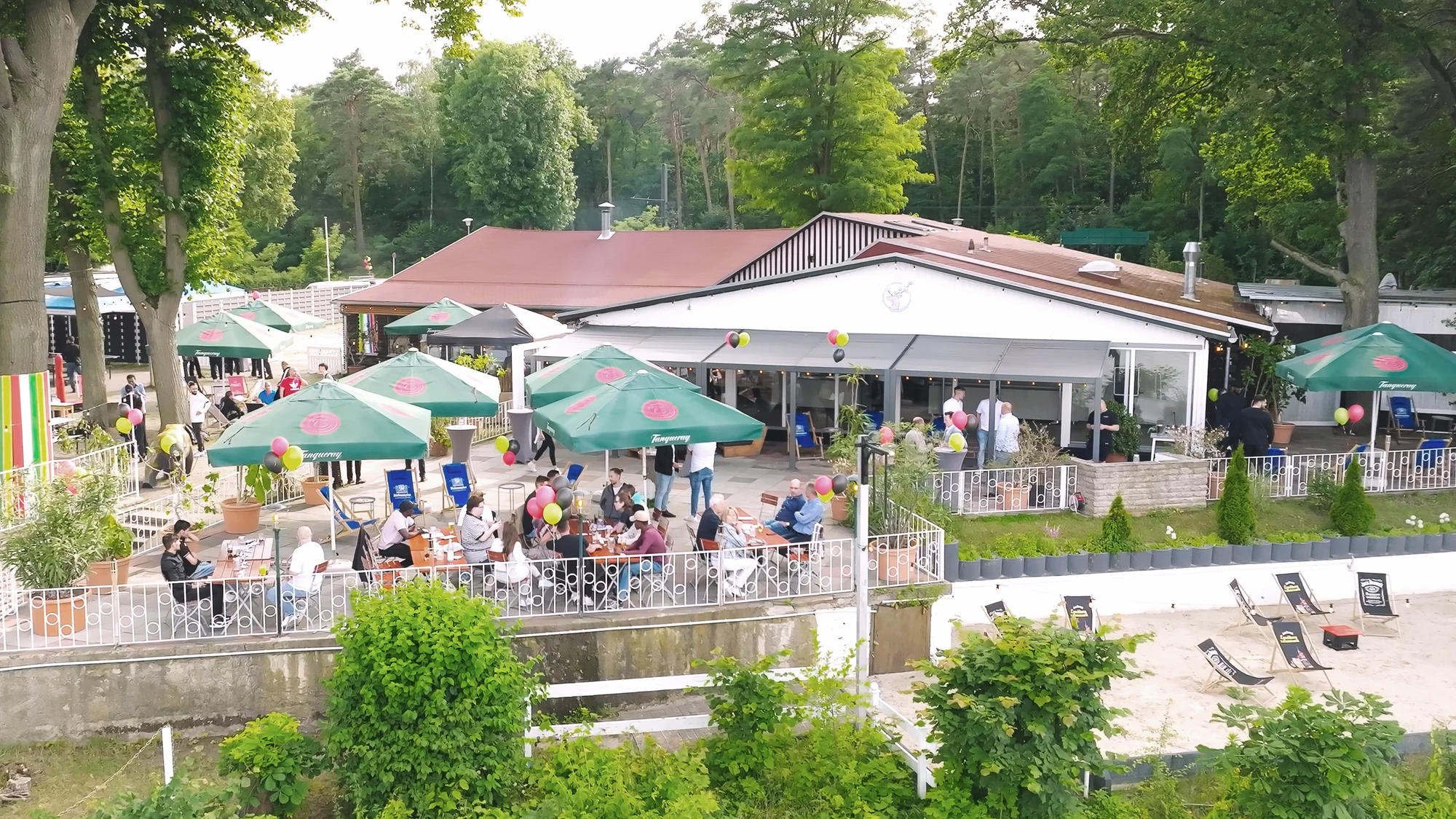 Sommerliche Strandterrasse mit vielen Gaesten, Schirmen und Seezugang.