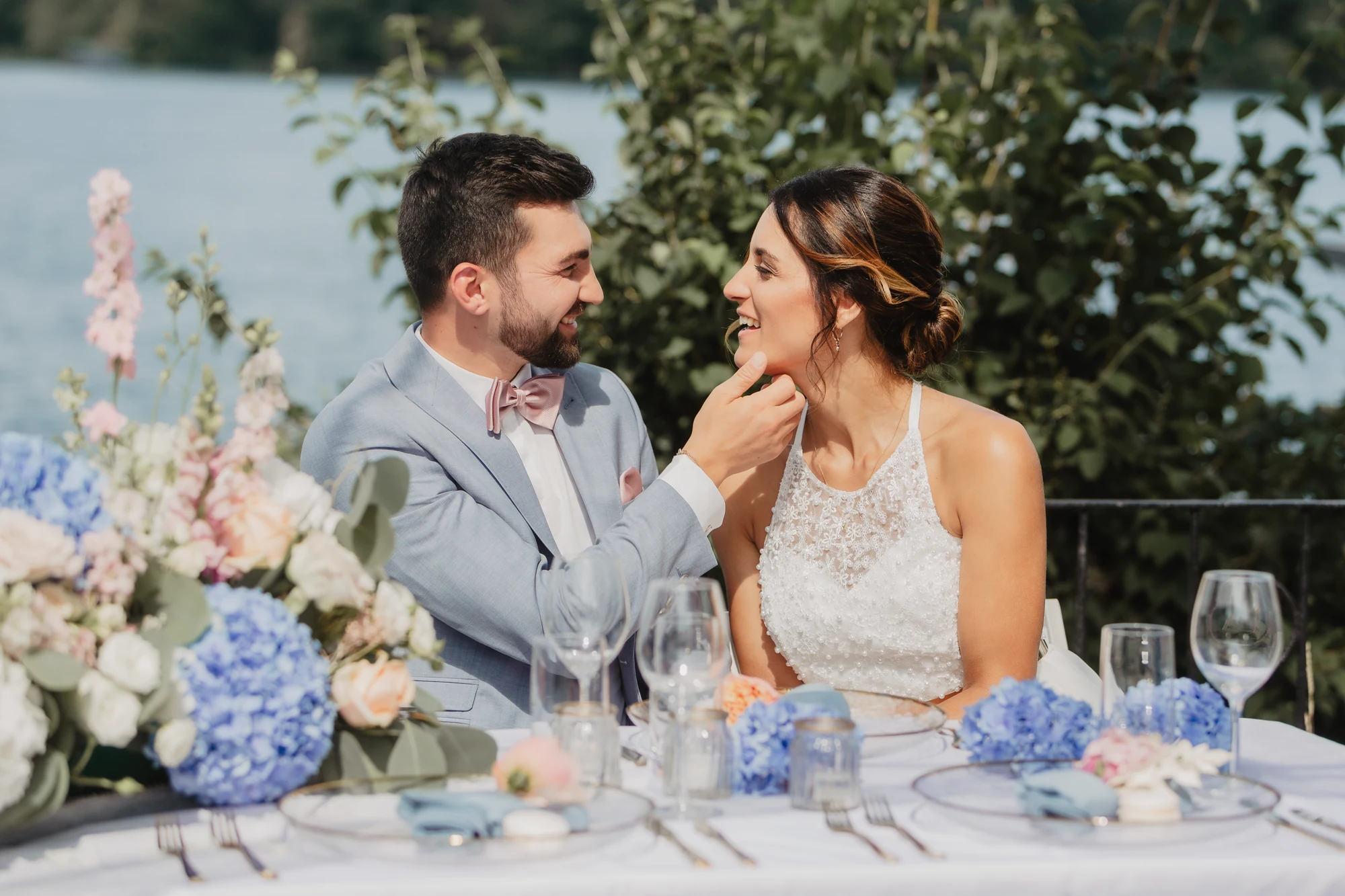 Brautpaar sitzt an einer gedeckten Tafel mit Blumen und Gruen im Hintergrund.