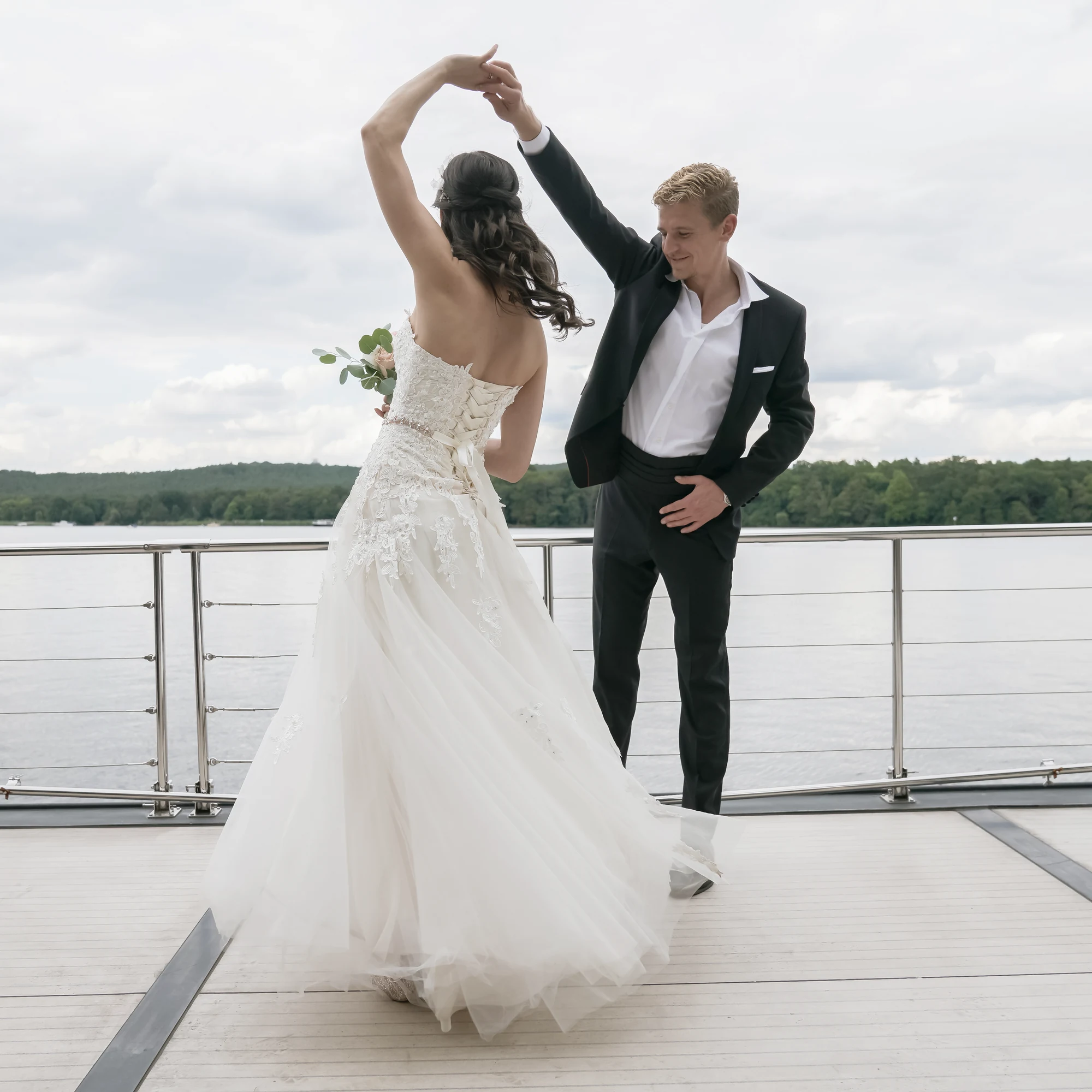 Brautpaar tanzt auf einem Hausboot vor grauem Himmel und weitem See.
