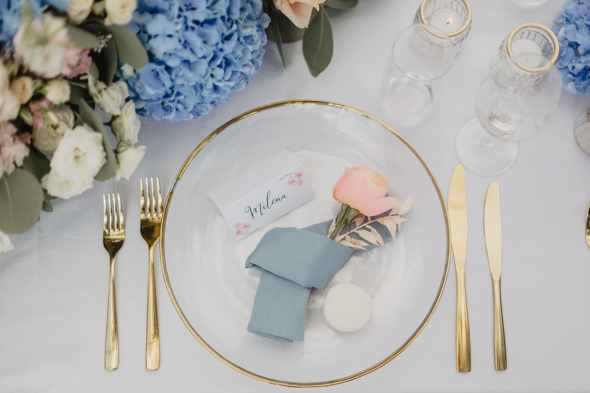Gedeckter Teller mit Serviette, Goldbesteck und Blumen auf weisser Hochzeitstafel.