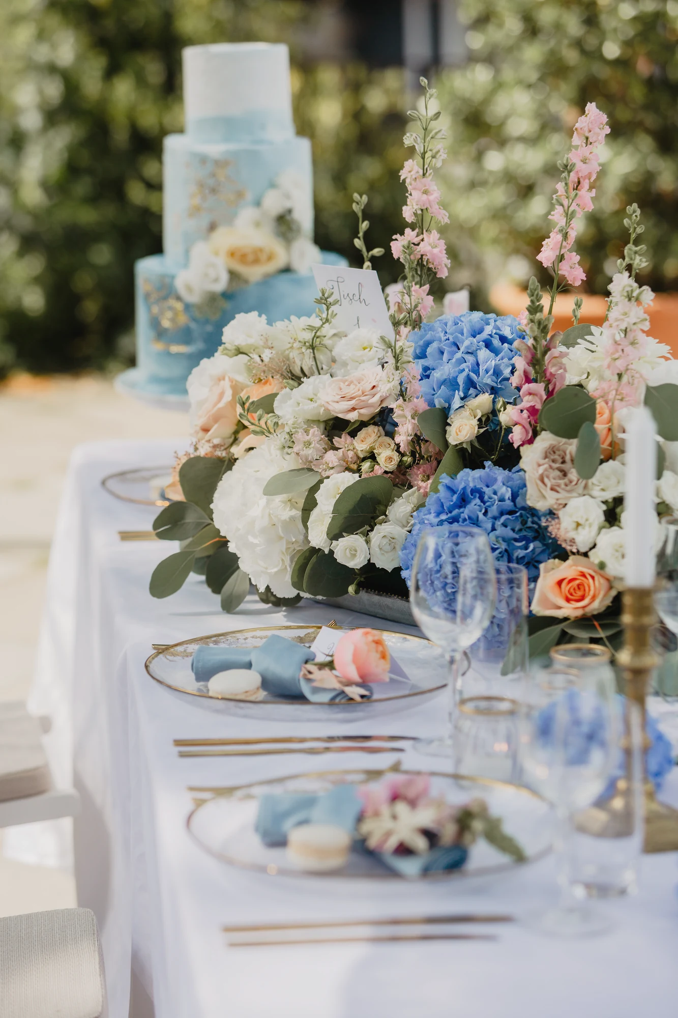 Lange Tafel mit blauem Gedeck, Blumen und Hochzeitstorte im Hintergrund.