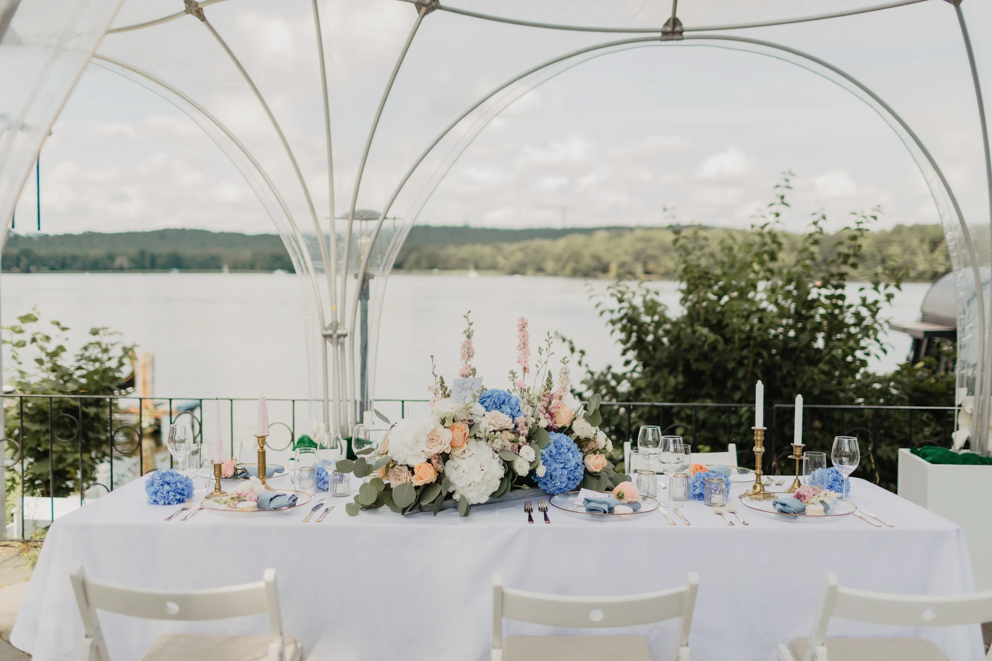 Gedeckte Hochzeitstafel unter Kuppelzelt mit Blumenarrangements am See.