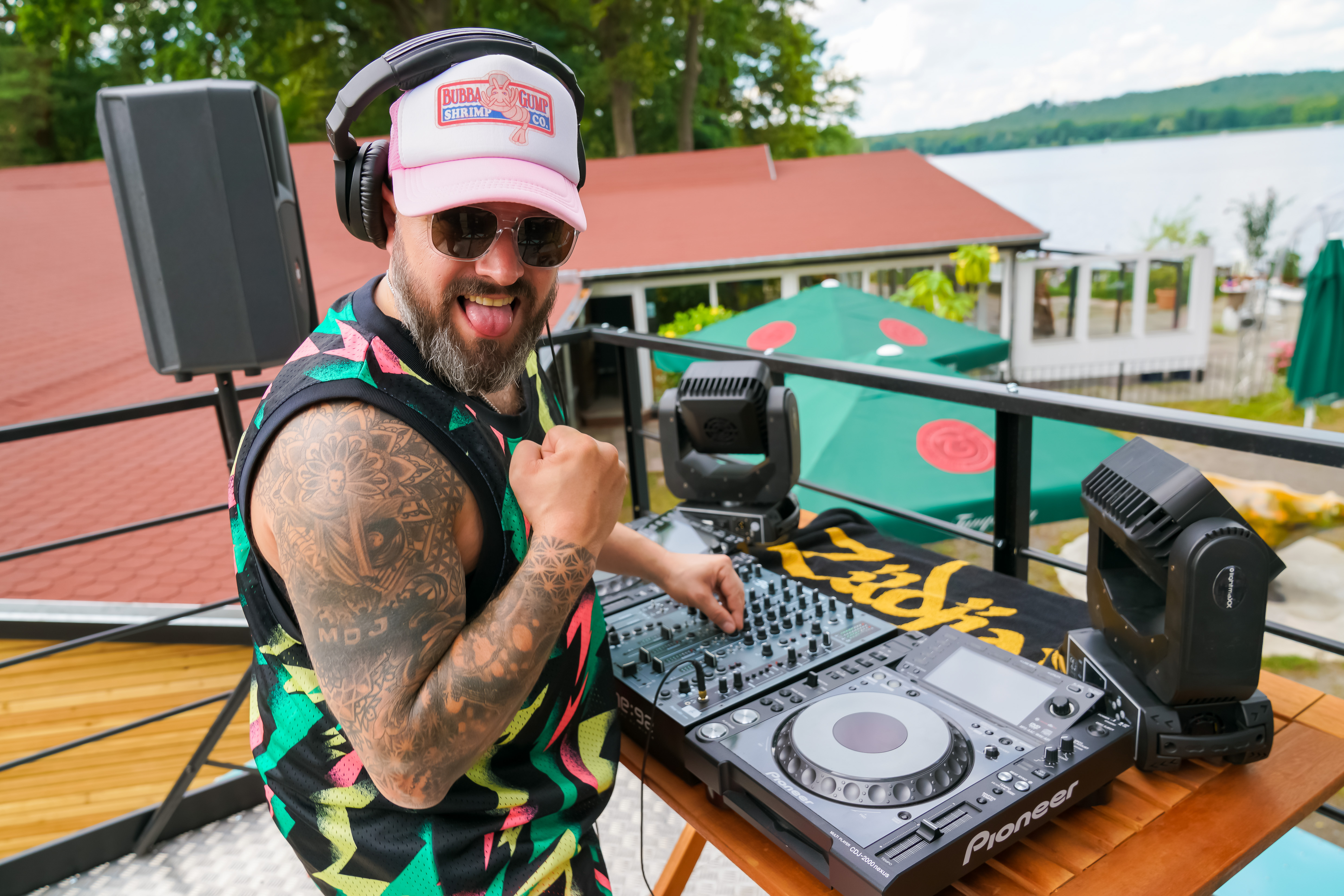 DJ steht auf einem bunten Truck mit Technik, See und Terrasse im Hintergrund.