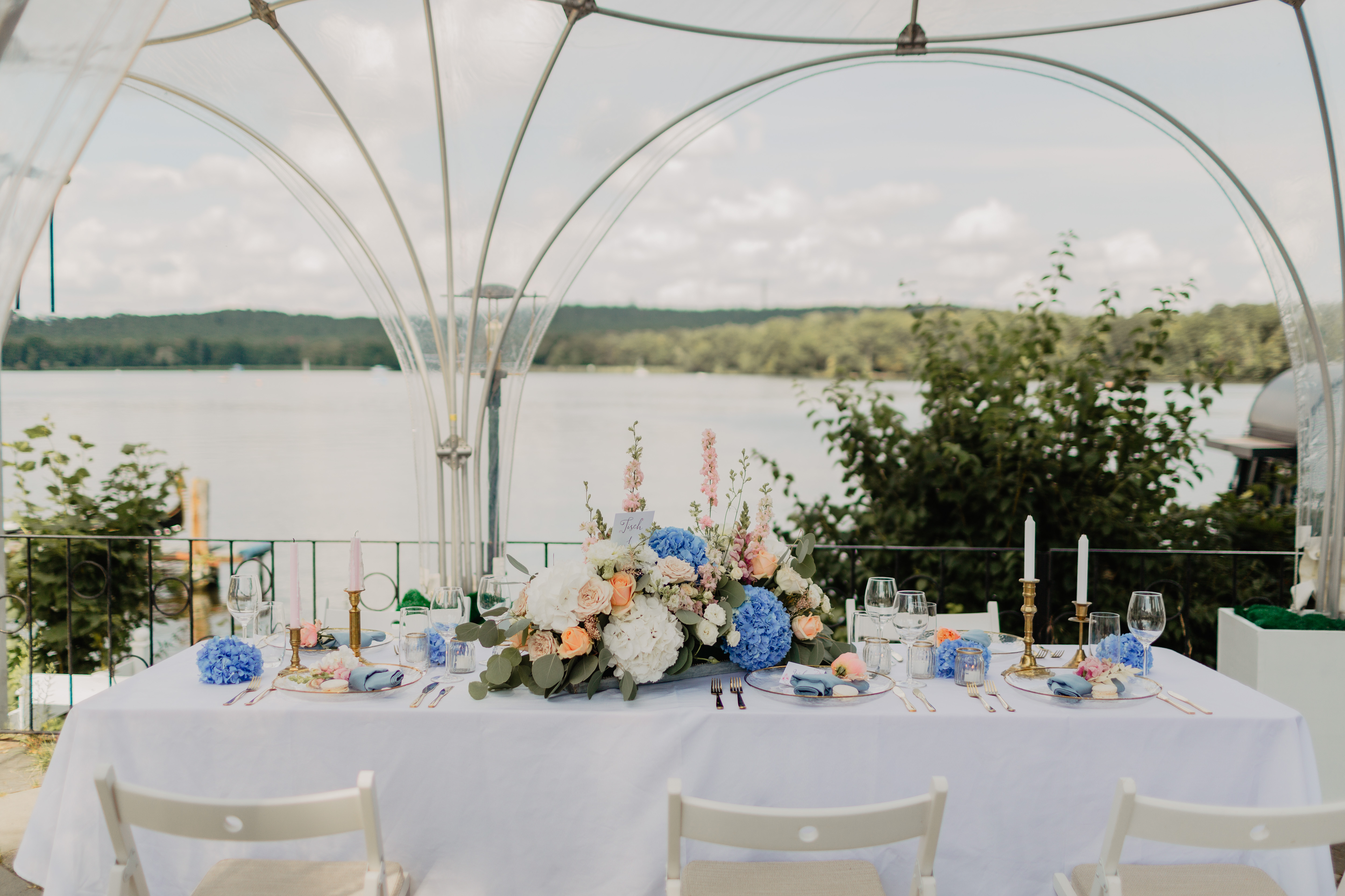 Gedeckte Hochzeitstafel unter Kuppelzelt mit Blumenarrangements am See.